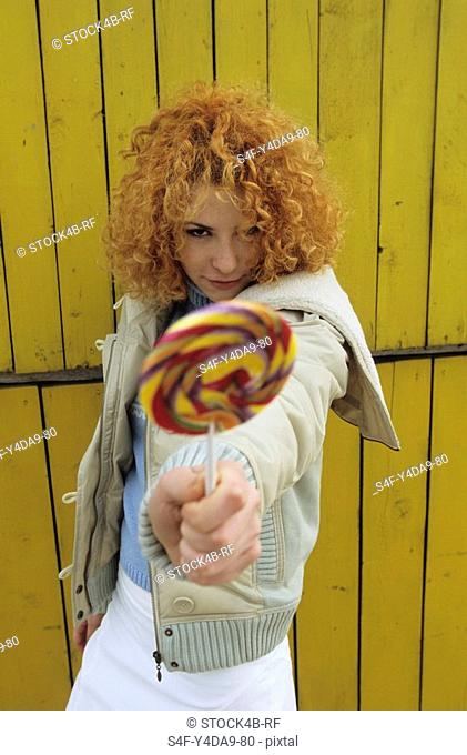 Young redhaired Woman elongating a Lollipop in the Face of the Spectator - Facial Expression - Allurement - Sweets