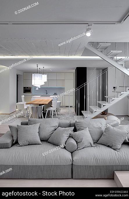 Interior in a modern style with white walls and a stair with metal railing. There is a gray sofa with pillows, kitchen zone with wooden table and chairs