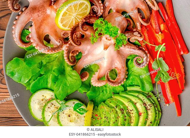 top down of octopus served with sliced avocado, lettuce, red pepper, lime, cucumber and sprig of pea leaves on plate, restaurant food