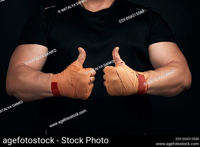 muscular athlete in black uniform shows a like symbol with two hands, palms wrapped in orange textile sports bandage, black background