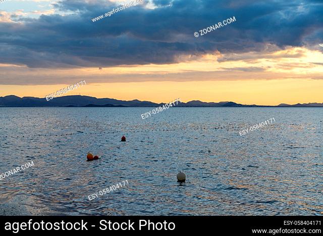 View of calm ocean waters under an expressive sky at sunset with mountain silhouette in the background and buoys in the foreground