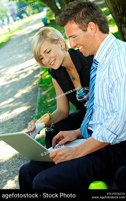 Young businesspeople sitting on bench in park having luch and looking at laptop computer