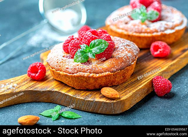 Delicious raspberry mini tarts (tartlets) with almond cream and green mint on a wooden serving board, selective focus