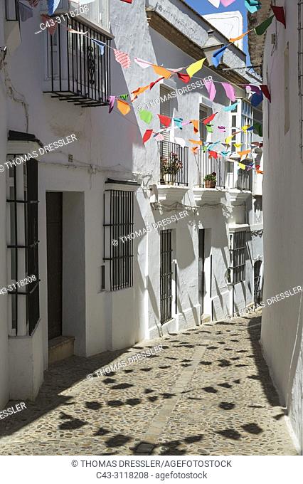 Alley and brilliantly whitewashed, reja (grille) fronted houses in the hilltop White Town of Arcos de la Frontera. The little colourful flags are remains of a...