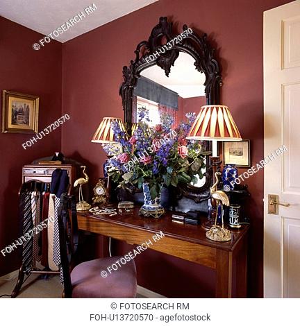 Carved mahogany mirror above antique table with lighted lamps and fresh floral arrangement in red bedroom