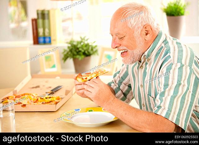 Smiling older man eating pizza slice sitting at living room table