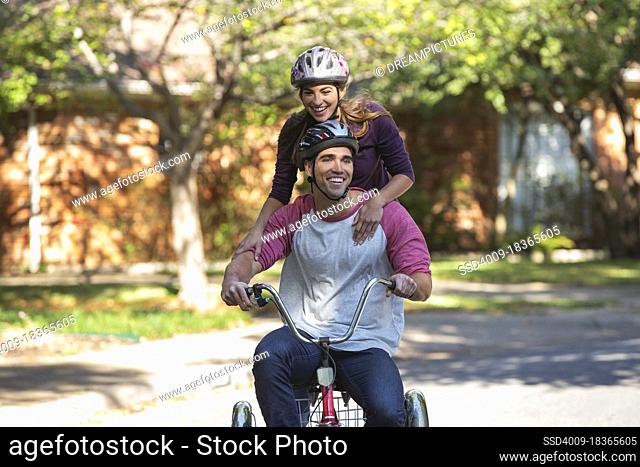 Young couple with helmets riding large tricycle through neighborhood, girl standing on back with her arms on boyfriends shoulders