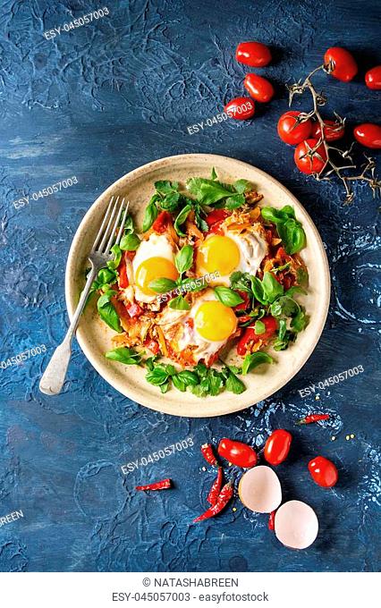 Traditional Israeli Cuisine dishes Shakshuka. Fried egg with vegetables tomatoes and paprika in ceramic plate with herbs and ingredients above over blue texture...