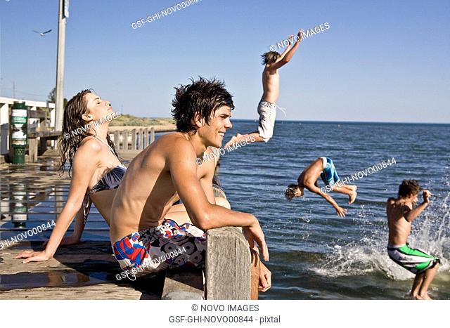 Young Couple Sitting on Edge of Pier With Group of Boys Jumping in Water