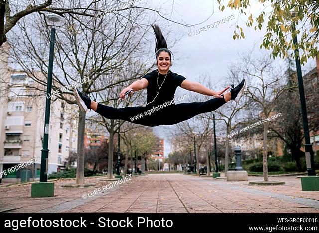 Portrait of young woman in black sportswear jumping in the air while listening music with headphones