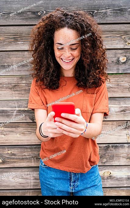 Smiling curly haired woman using smart phone in front of wooden wall