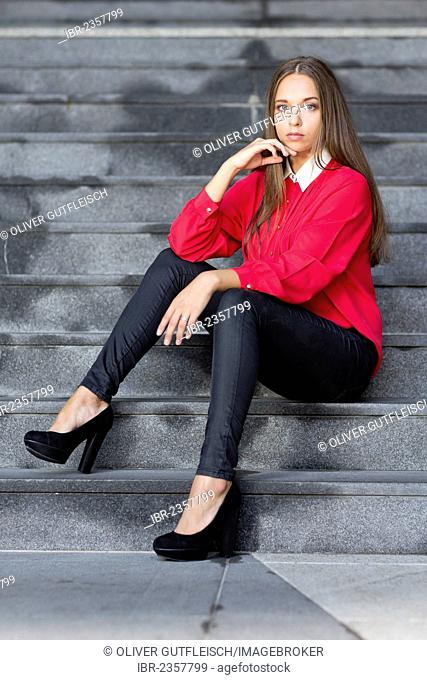 Young woman wearing a red top, black trousers and high black shoes posing while sitting on stairs