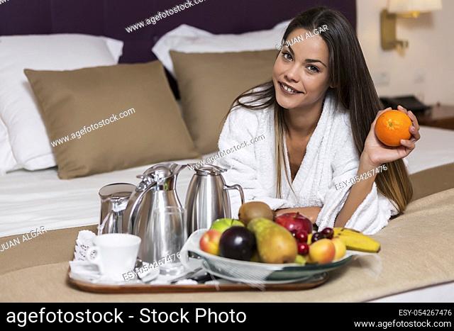 Charming brunette in white bathrobe lying on bed with plate of fruit and served tea on tray