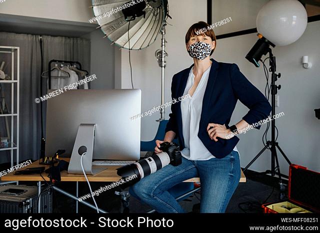 Female photographer sitting with camera on desk in studio during pandemic