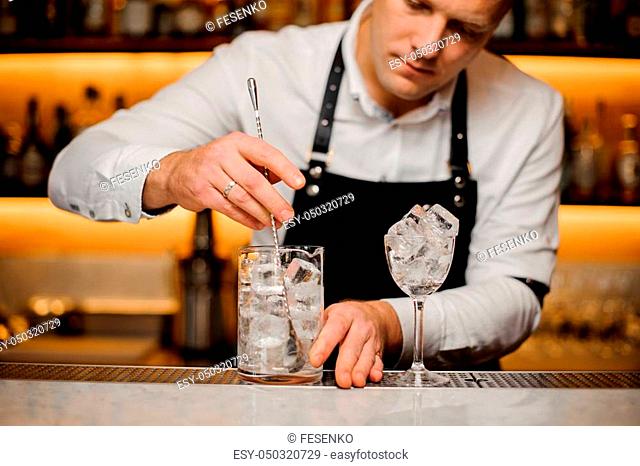 Young barman dressed in a white shirt and apron stirring ice cubes in a glass using a spoon