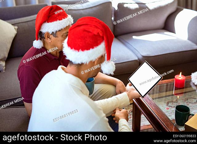 Biracial adult son and senior father in santa hats making tablet christmas video call