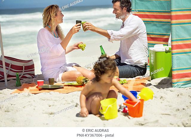 Couple toasting with drink while their daughter playing on the beach