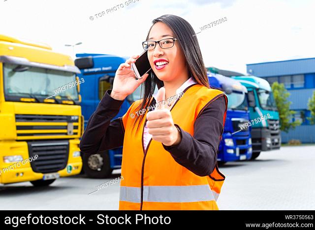 Logistics - female Asian forwarder or supervisor with mobile phone, in front of trucks and trailers, on transshipment point