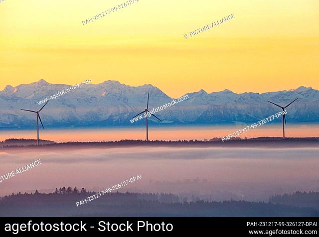 17 December 2023, Baden-Württemberg, Uttenweiler: Three wind turbines rise out of the fog in the morning shortly after sunrise, while the Alps