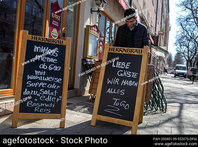 01 April 2020, Bavaria, Munich: An Indian cook puts up signs in front of his restaurant in the Haidhausen district. On one of them is written: ""Dear guests