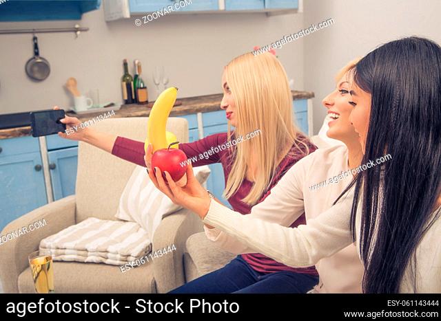 Toned picture of friends girls posing for mobile or smart phone's camera with different fruits while having party at home. Home party concept