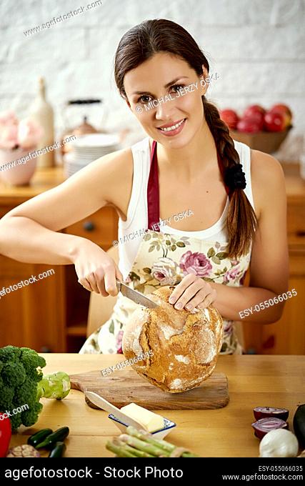 beautiful young woman, brunette slicing bread in the kitchen at a table full of organic vegetables