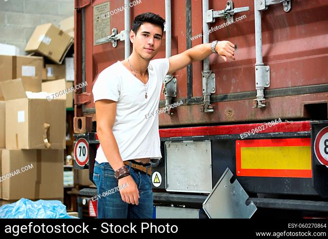 Waist Up Portrait of Young Smiling Man Next to Freight Truck Leaning against it, Looking at Camera