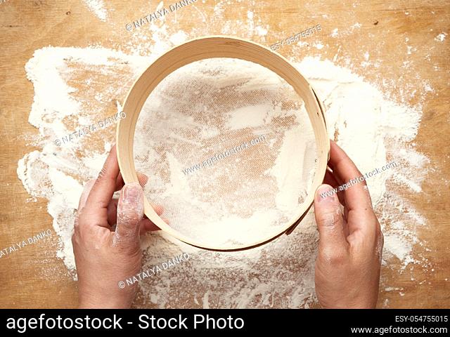 two female hands hold a round wooden sieve over a table with white flour, top view