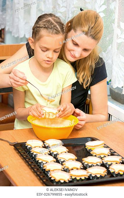 Mother helps daughter to pour batter into the mold for cupcakes