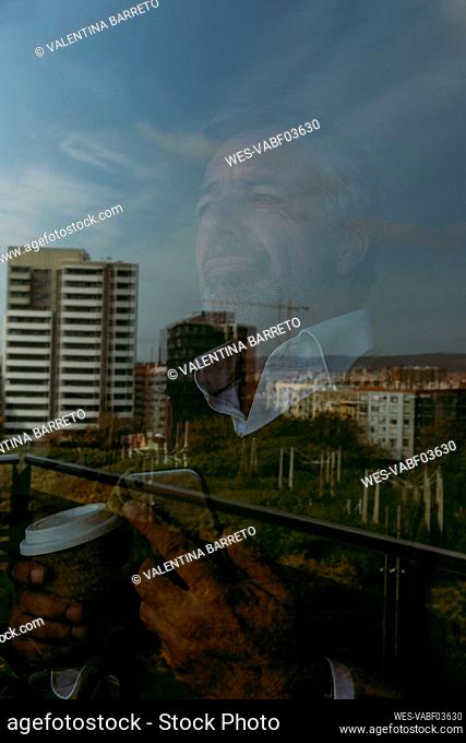 Reflection of man with coffee cup and mobile phone admiring city view on window glass at home
