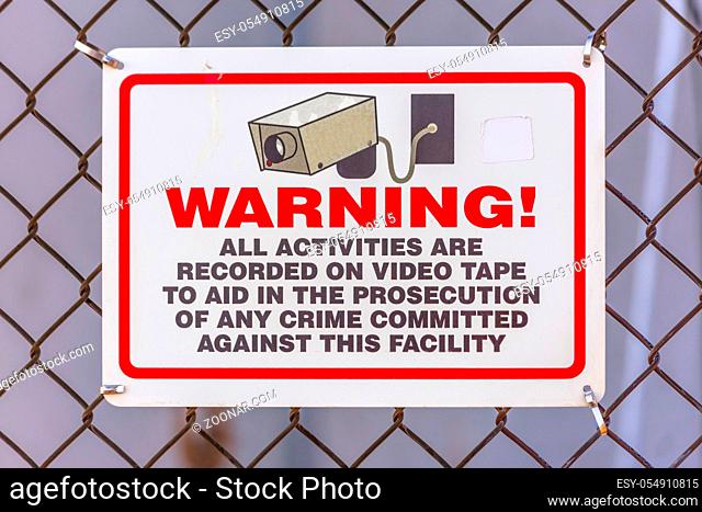 Warning security camera sign on a chain link fence. The sign says all activities are recorded on video tape to aid in the prosecution of any crime committed...