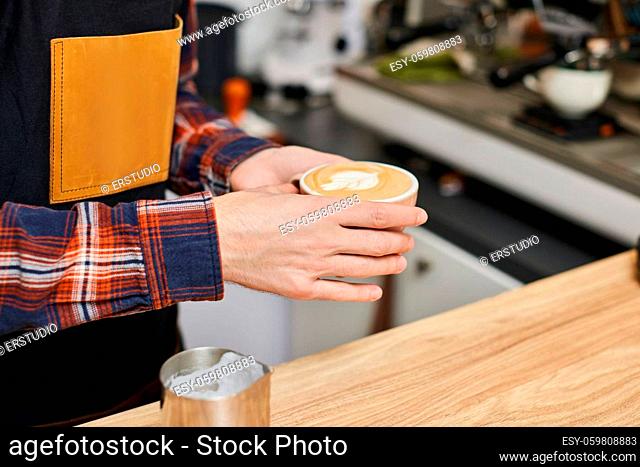 male waiter in apron holding cup of hot drink in coffee shop, close-up