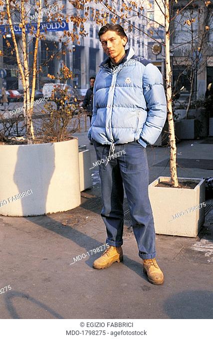 A young guy, portrayed in a serious attitude in Piazza San Babila, wears a Moncler jacket, blue jeans and Timberland boots; he's a paninaro