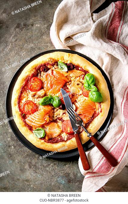 Traditional pizza with smoked salmon, cheese, tomatoes and basil served on black plate with kitchen towel over old dark metal background