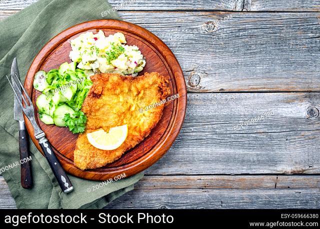 Traditional deep-fried schnitzel with potato and cucumber salad offered as top view on a rustic wooden board with copy space right