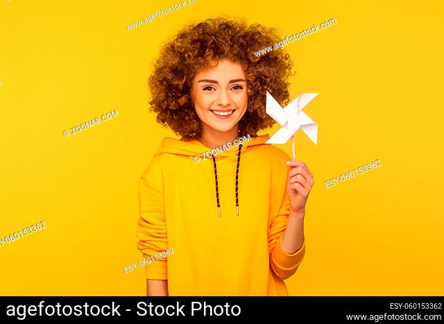 Origami hand mill. Portrait of happy curly-haired woman in urban style hoodie smiling carefree and holding paper windmill, pinwheel toy on stick