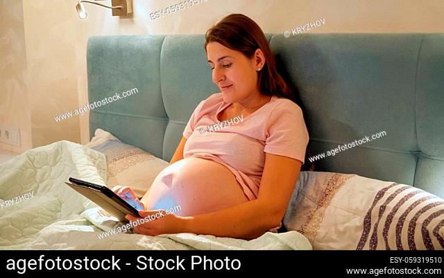 Smiling pregnant woman wearing pajamas working on tablet and browsing internet before going to sleep in bed at night