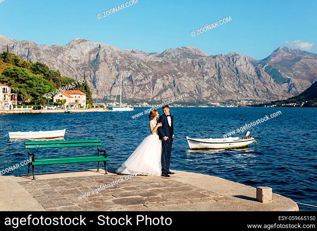 The bride and groom are standing ang hugging on the pier near the old town of Perast near green bench, small boats behind them . High quality photo