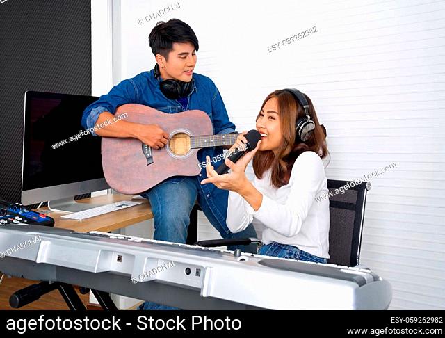 Young asian woman sings in front of white wall. Her boyfriend play the guitar together. Musicians producing music in professional recording studio