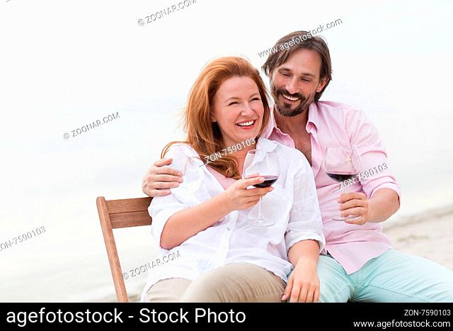 Couple embracing each other#39;s at teh beach. People in love smiling, drinking red wine from glasses and sitting on comfortable chairs