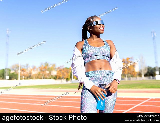 Happy female athlete wearing sunglasses looking away while holding energy drink bottle on sports track