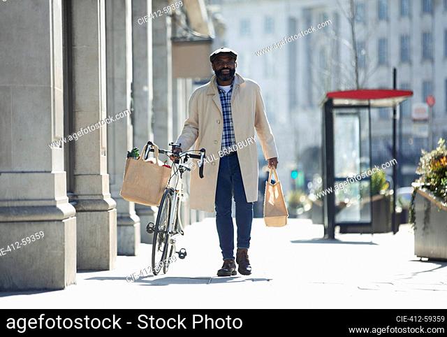 Man with. bicycle and groceries walking on sunny city sidewalk