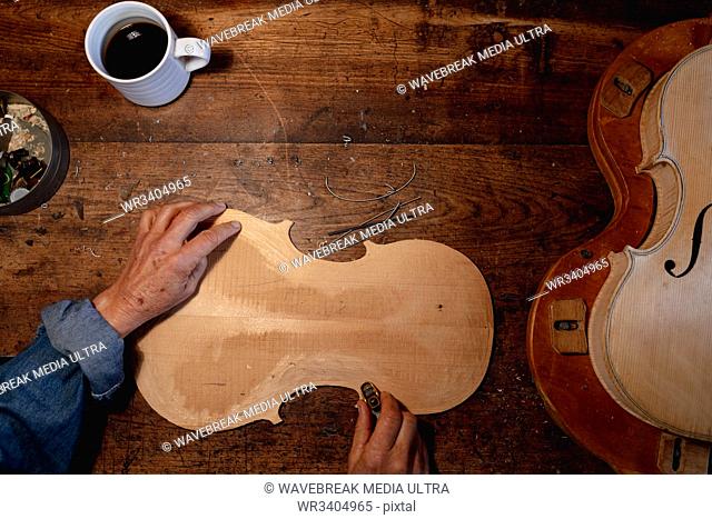 Overhead view of a senior Caucasian female luthier working on the body of a violin in her workshop, with coffee cup on a workbench