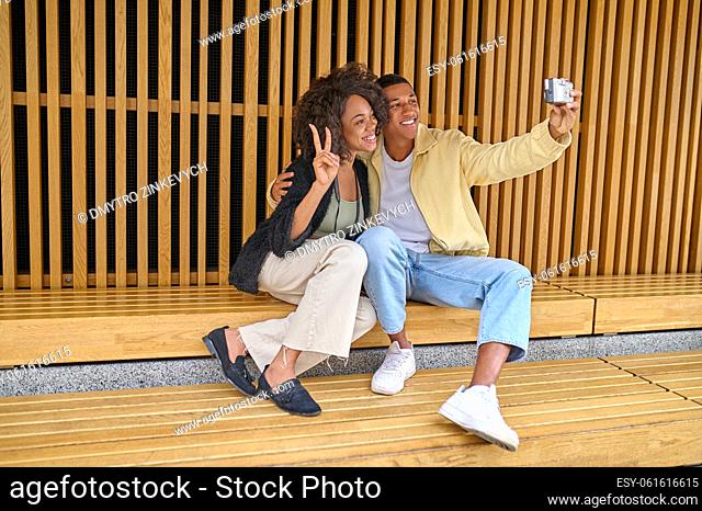 Selfie. Smiling dark-skinned guy hugging girl showing freedom sign taking selfie on camera sitting outdoors on fine day