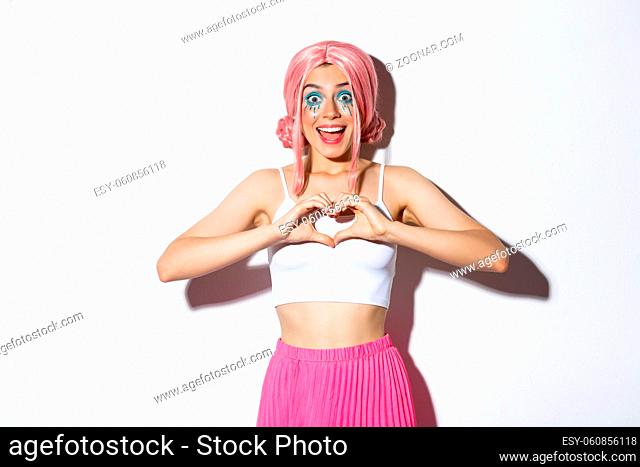 Image of excited smiling girl like something, showing heart gesture and looking amazed, wearing halloween costume with bright makeup and pink wig