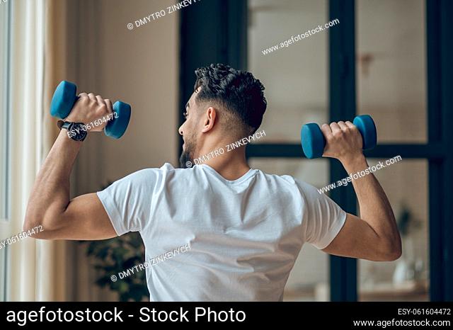 Keeping fit. Young dark-haired man exercising with dumbbells and looking contented