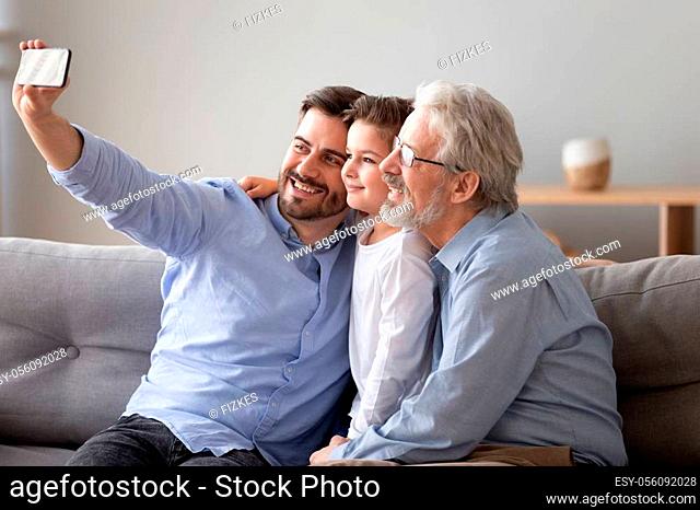 Three generations relatives people men sitting on couch in living room, grandad grown up son grandkid using smart phone take selfie photo smiling feels happy...