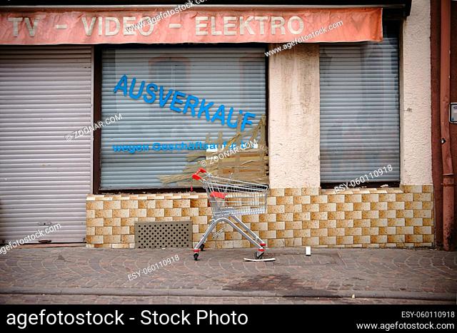 Ein leerer Einkaufswagen steht auf dem Gehweg vor einem wegen Geschäftsaufgabe geschlossenen Elektrowarengeschäftes mit eingebrochener Fensterscheibe