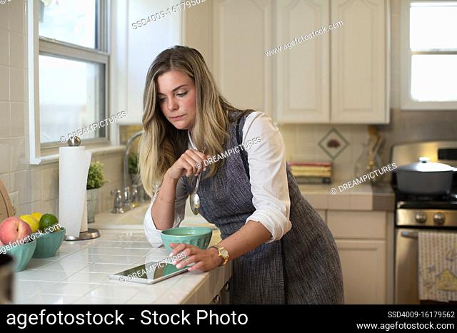 Young woman in her kitchen, eating soup while using her iPad