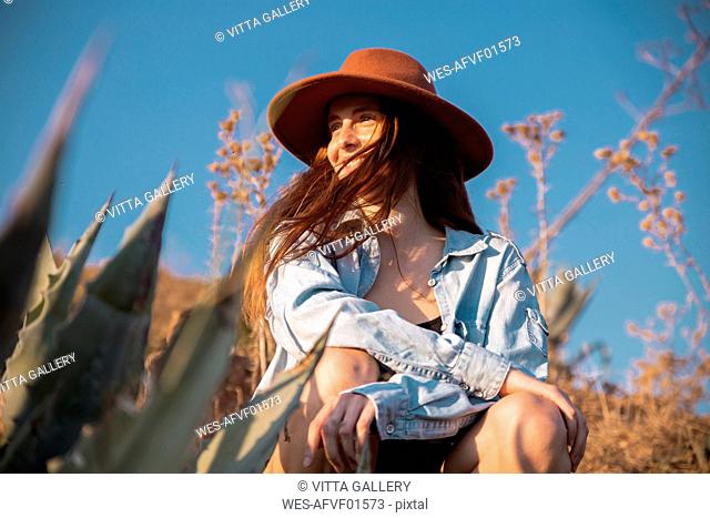Smiling young woman wearing a hat sitting at an agave in the countyside